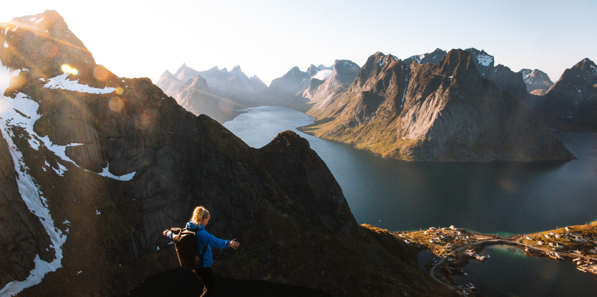 A woman standing on a mountain with a fjord and snow capped peaks in the background
