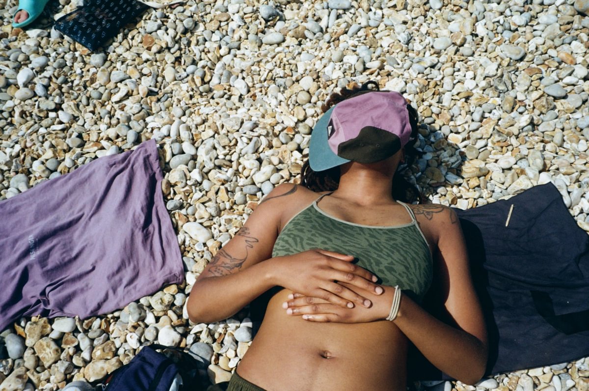 A person lies on a pebble beach wearing a green sports bra, a pink and blue visor, and a black top, with purple and black towels beside them.