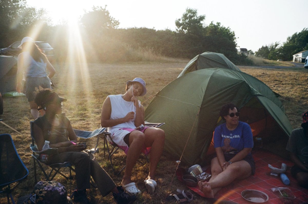 A group of people sitting around a green camping tent at an outdoor campsite during late afternoon sunlit hours.