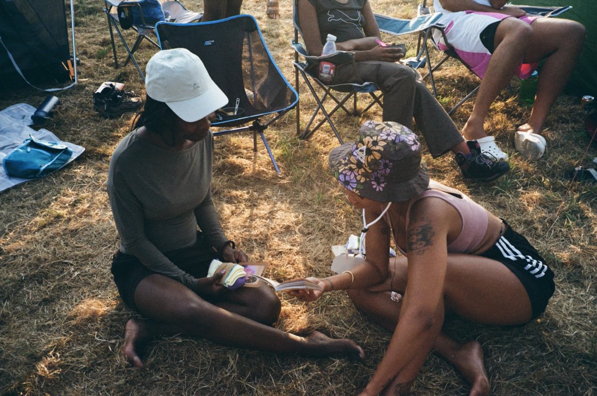 Two people sitting on the grass at a campsite, with folding chairs and other people visible in the background. One person wears a white baseball cap, a gray long-sleeve top, and black shorts, while the other wears a floral bucket hat, a pink tank top, and black shorts with white stripes.