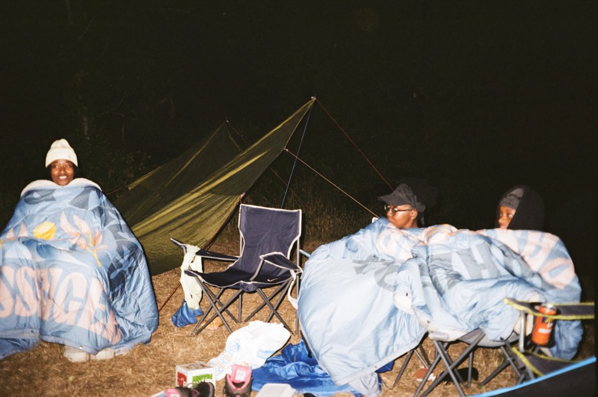 Three people sitting in camping chairs wrapped in patterned blankets at night in front of a tent, with a folding chair in the center and camping gear on the ground.