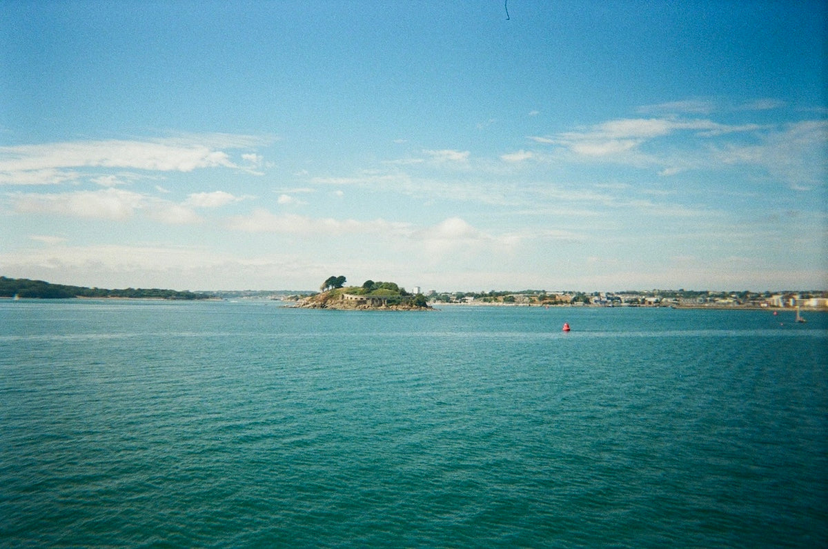 A scenic photograph of a calm blue-green body of water under a bright blue sky with light clouds, featuring a small rocky island with trees in the center and distant shoreline buildings on the horizon.