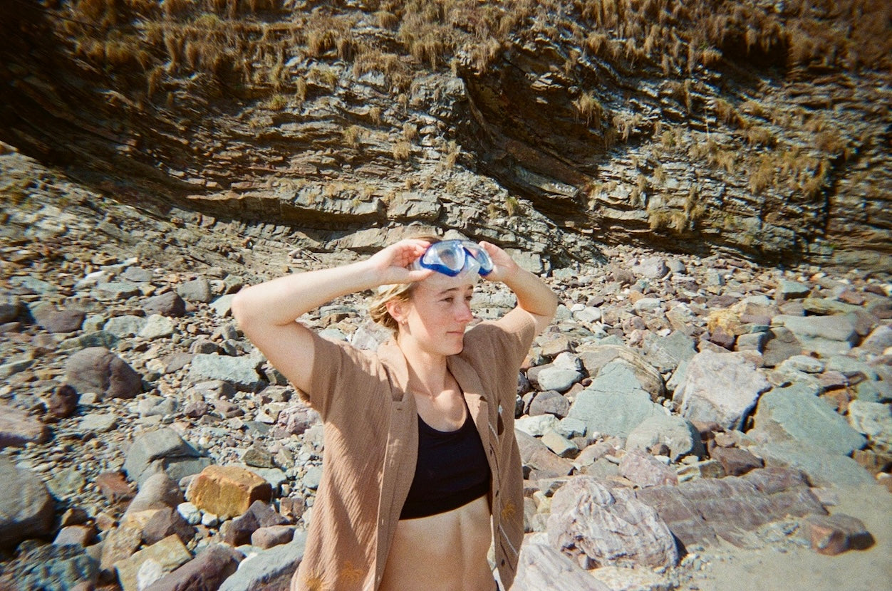 A woman standing on a rocky beach or cliffside, adjusting blue swim goggles on her head. She is wearing a light brown linen shirt over a black tank top, with a rugged stone wall and dry grass in the background.