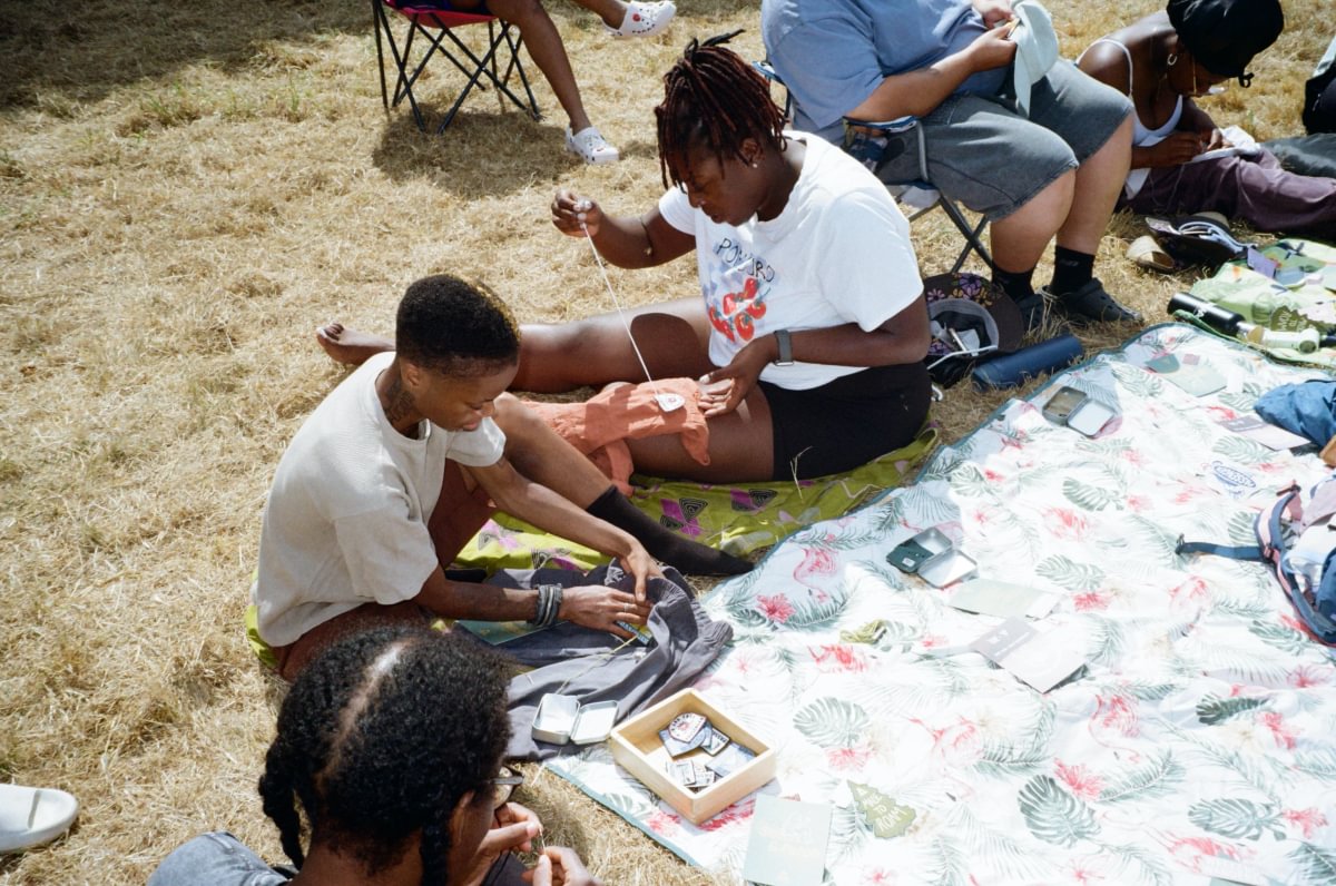 Several people sitting on a grassy lawn outdoors, working on or arranging colorful fabric pieces on a patterned blanket. The scene is casual and informal, with folding chairs and dry grass visible.