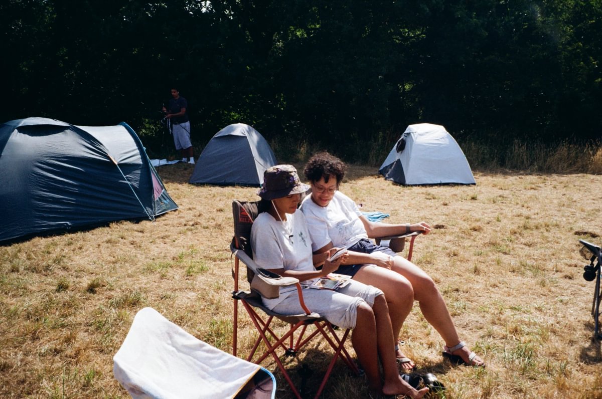 Two people sitting in folding chairs at a campsite, with three camping tents pitched on dry grass in the background among trees.