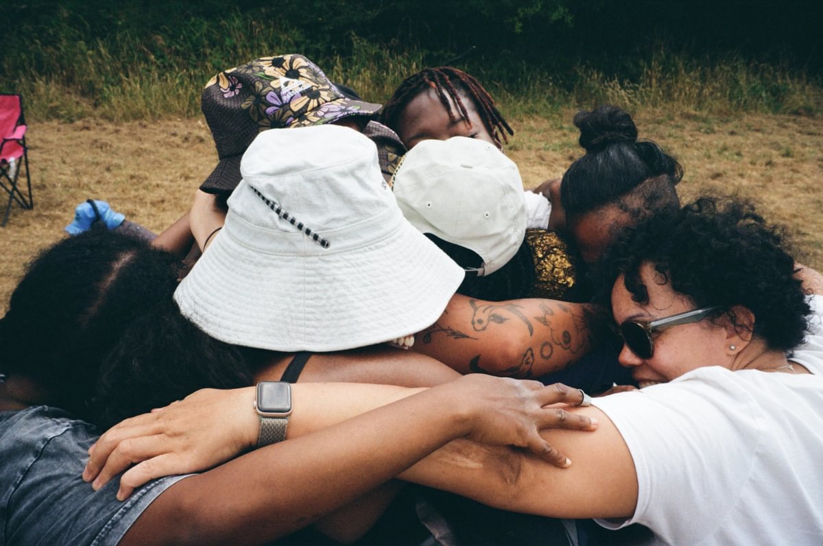 Several people huddled together outdoors, with one person wearing a large white bucket hat and another wearing a white baseball cap. The group is standing on grass with trees in the background.