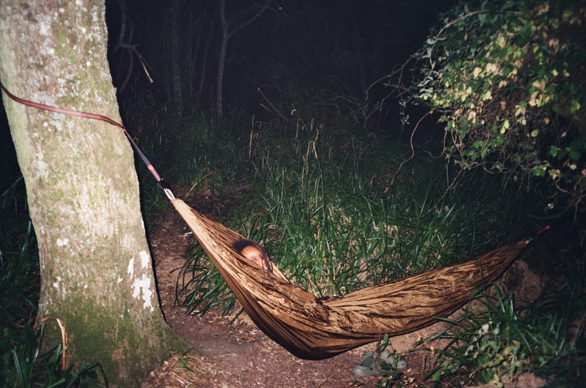 A brown hammock strung between a tree and a bush, with a person sleeping inside, photographed at night in an outdoor forest setting.