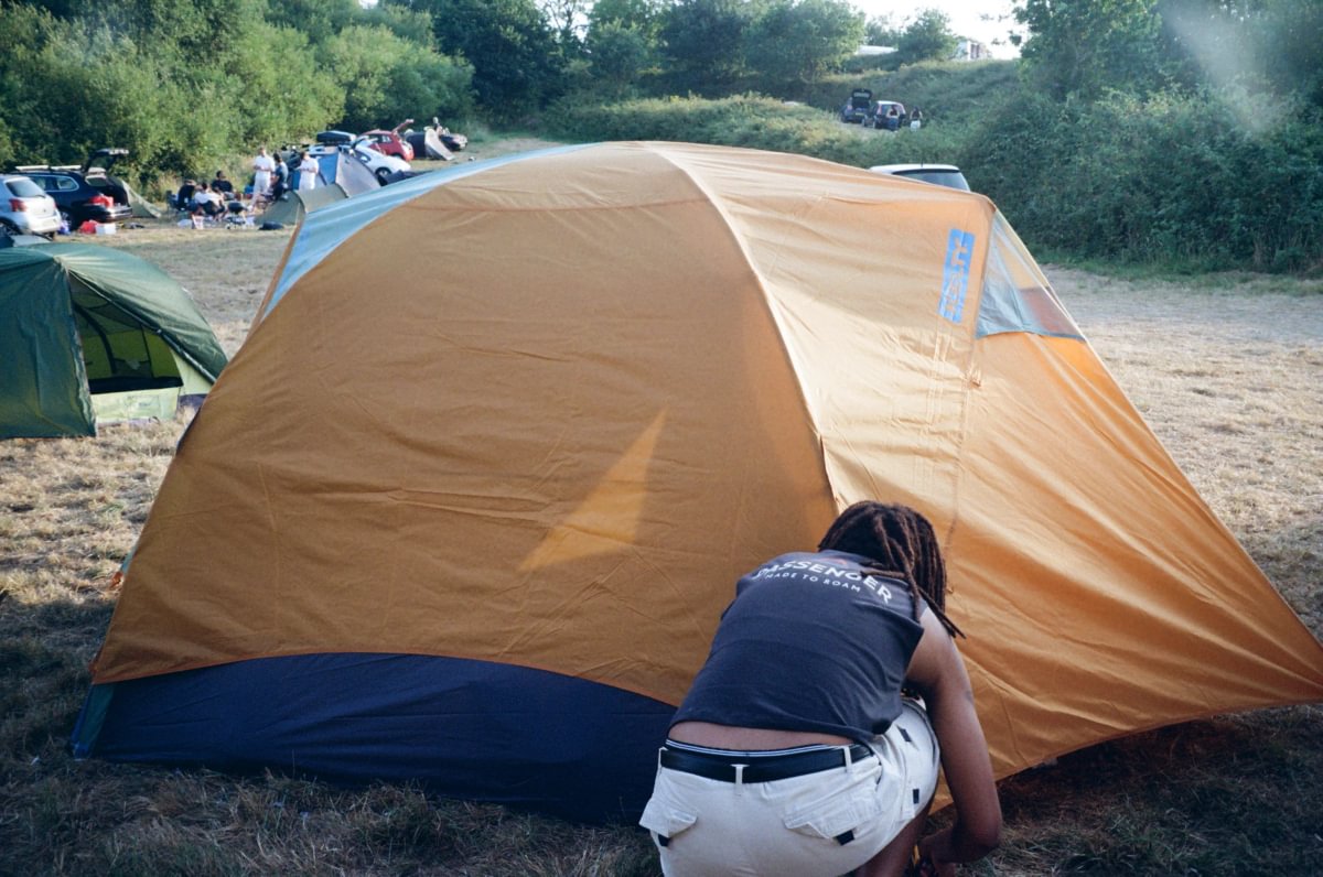 A person crouches in front of a large camping tent pitched outdoors on grassy ground, with trees, other tents, and campsite activity in the background.