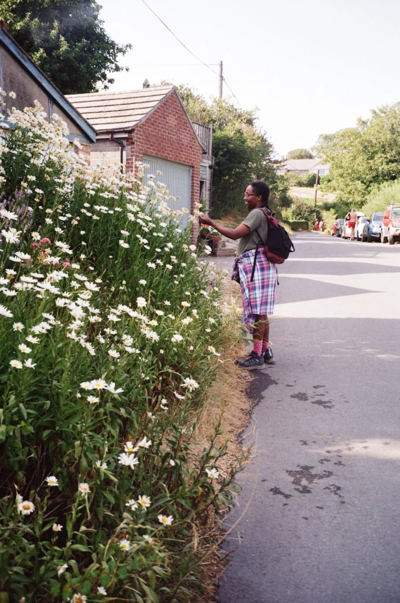 A person standing on a street beside a large bush of white flowers, wearing a plaid skirt, green top, and backpack, with houses and trees in the background.