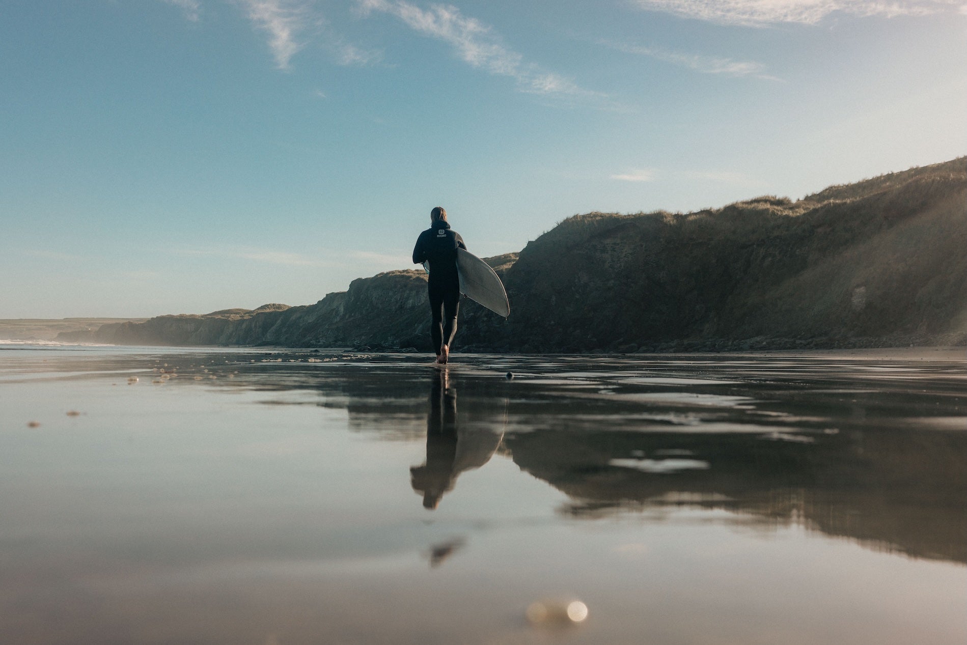 Person holding a surfboard walking on a beach with cliffs in the background