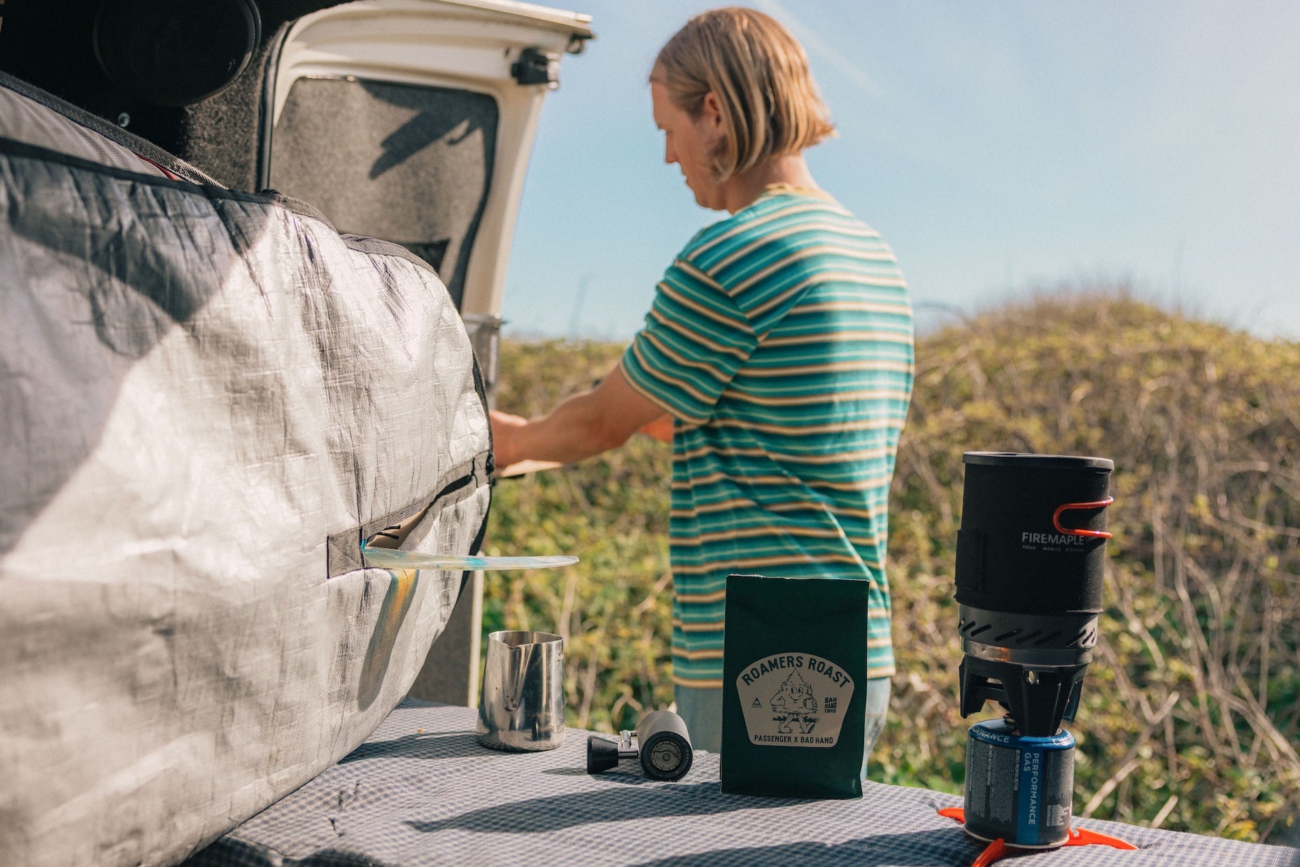 Person standing next to an open car trunk with camping gear, including a coffee press and a thermos.